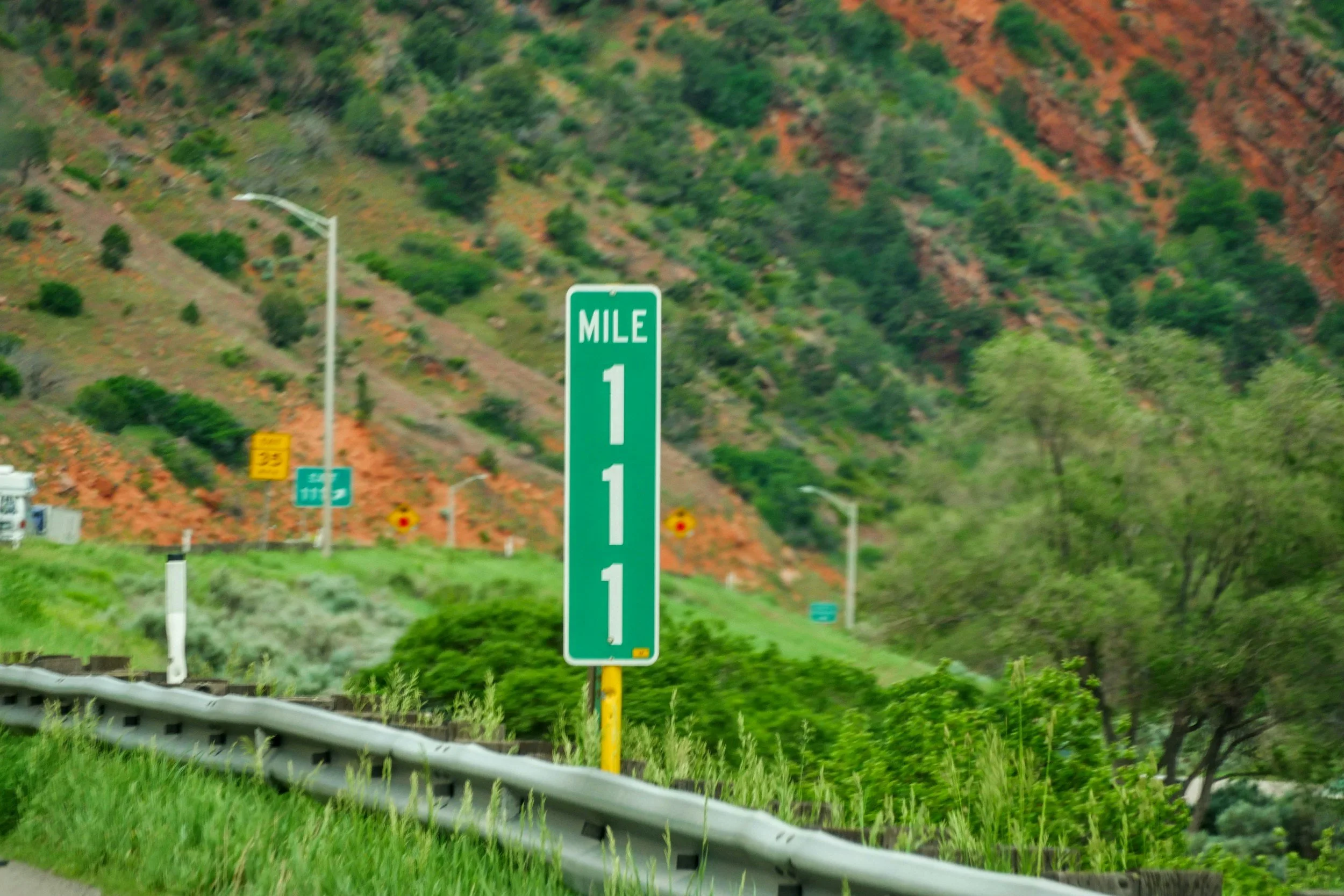 Highway sign indicating the first mile of a highway, with a scenic hillside covered in green trees and reddish soil in the background.