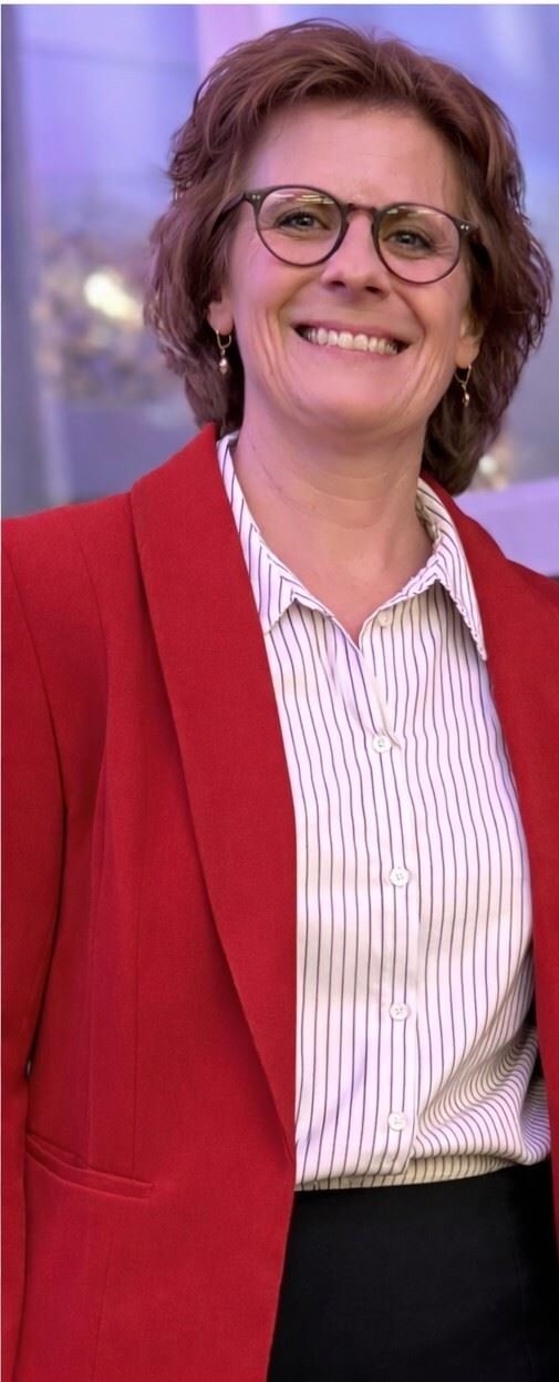 A smiling woman with glasses, brown hair, wearing a red blazer over a striped white shirt, standing indoors.