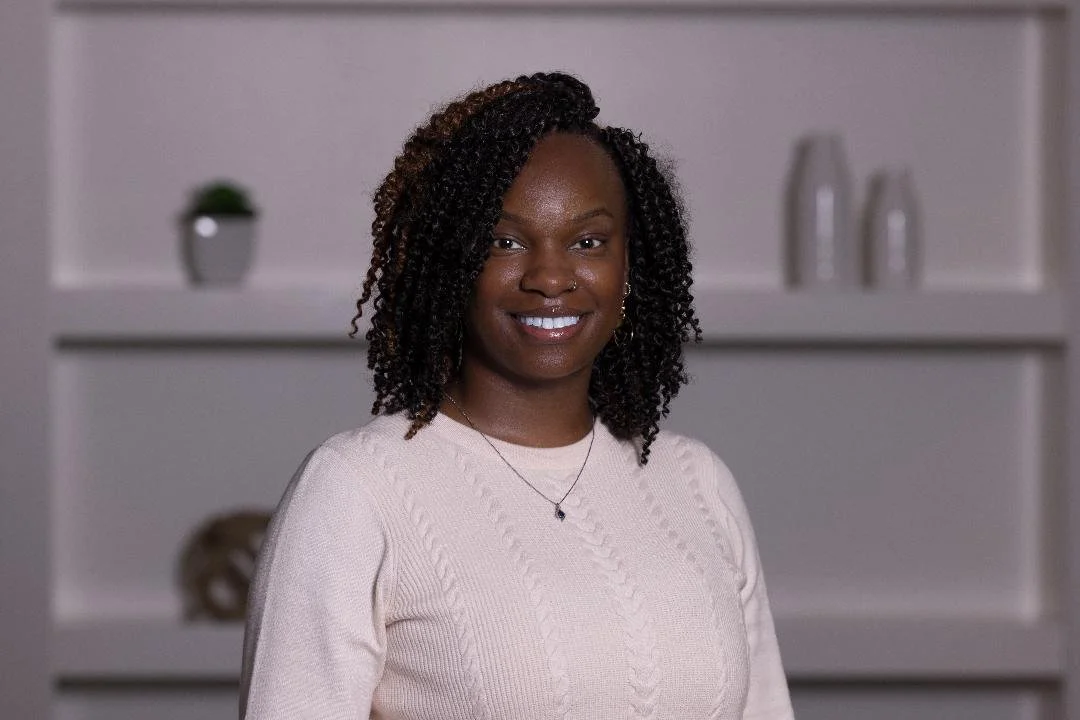 A smiling Black woman with curly hair wearing a light pink sweater and a necklace, standing in a room with white shelves and decorations in the background.
