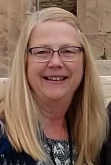 A woman with blonde hair, glasses, and a smile, standing outdoors with a stone wall in the background.