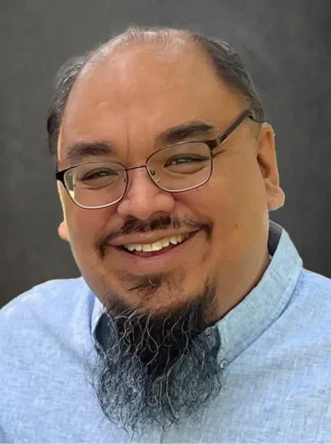 A man with glasses, a beard, and a mustache smiling, wearing a light blue shirt.