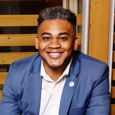 Smiling young man in a blue suit and white shirt, sitting indoors with wooden decor in the background.