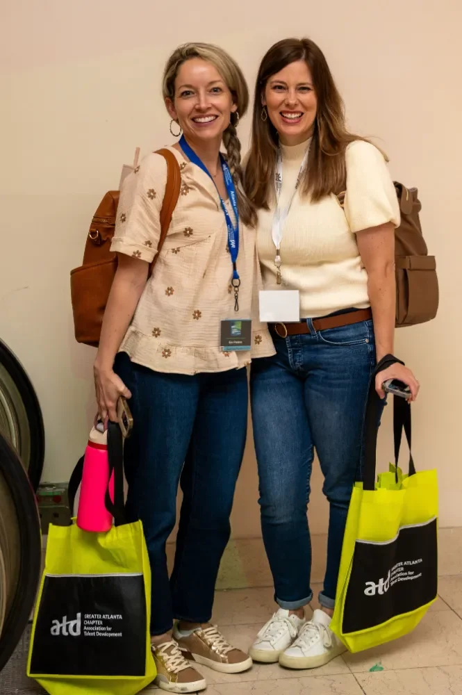 Two women smiling at the camera, standing in front of a plain wall, each holding a bright yellow tote bag with black accents and a logo, wearing casual clothes with conference badges around their necks.