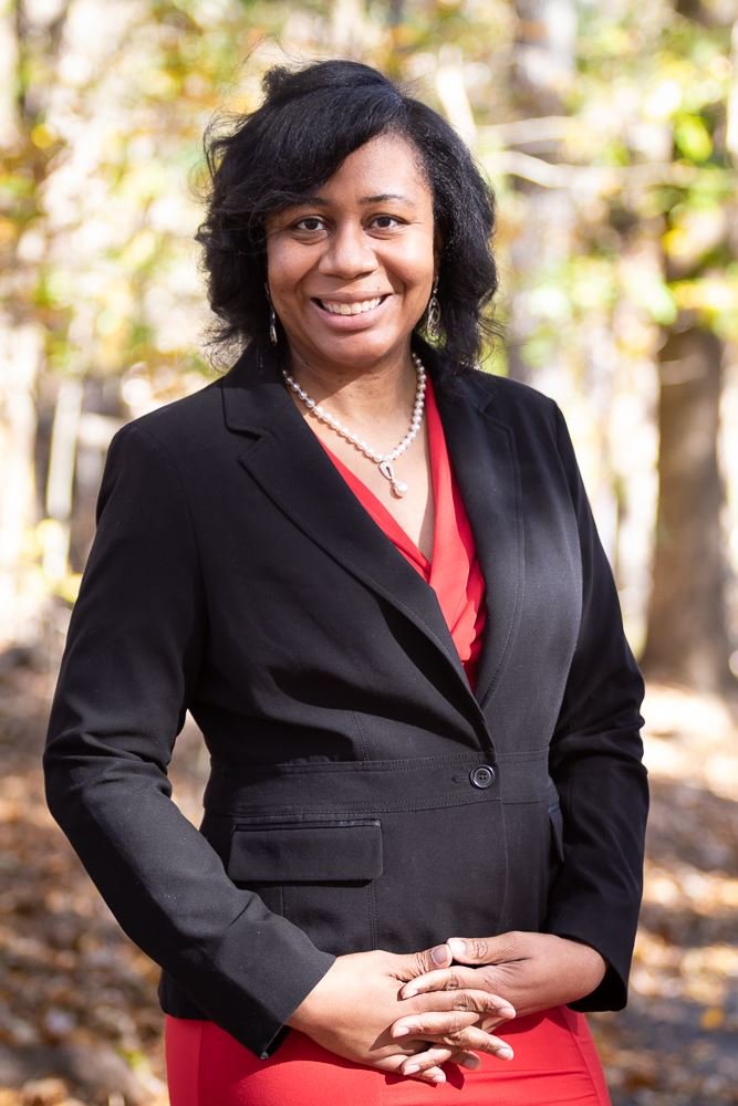 A woman standing outdoors among trees, wearing a black blazer over a red dress, with a pearl necklace and earrings, smiling at the camera.