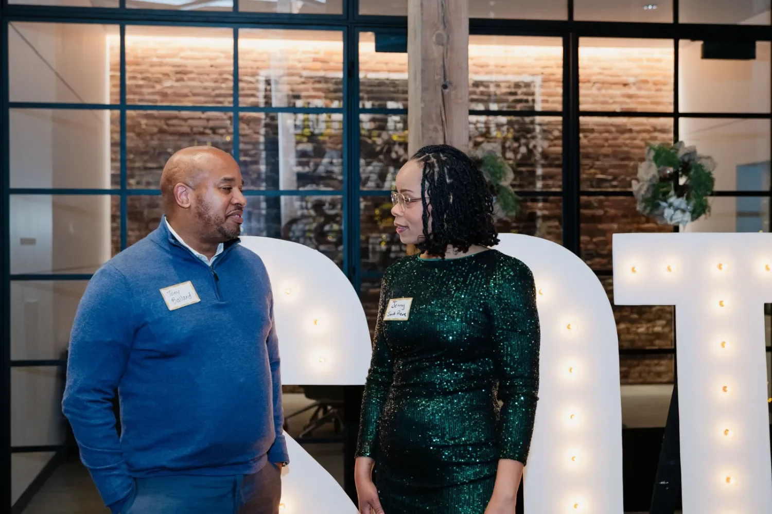 A man and woman are having a conversation in a modern, industrial-style space with large white letter lights in the background, decorated with greenery. Both are wearing name tags.