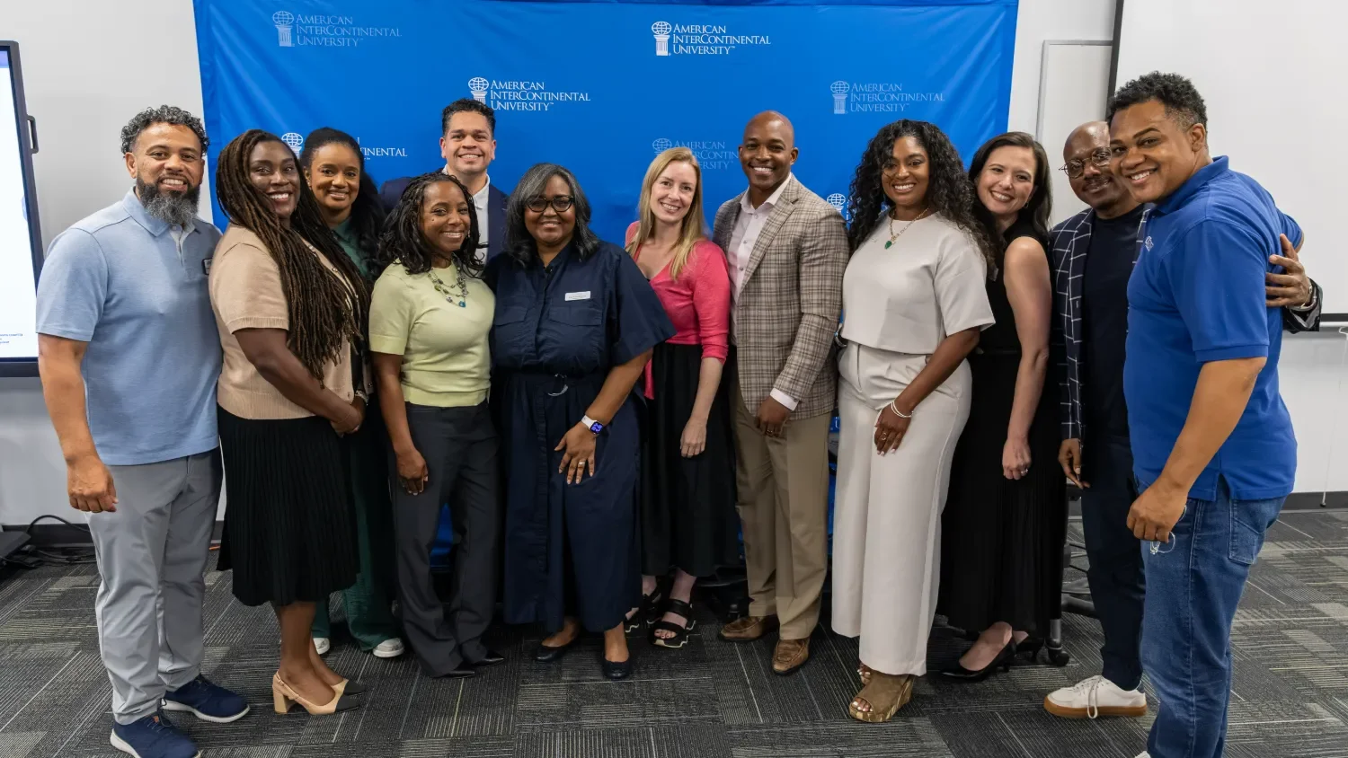 Group of diverse people posing together in front of a blue American Intercontinental University backdrop in a conference room.