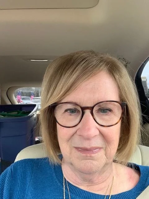 A woman with shoulder-length blonde hair wearing glasses and a blue top, sitting in a car, with the back of the car's interior and a parking lot visible through the window.