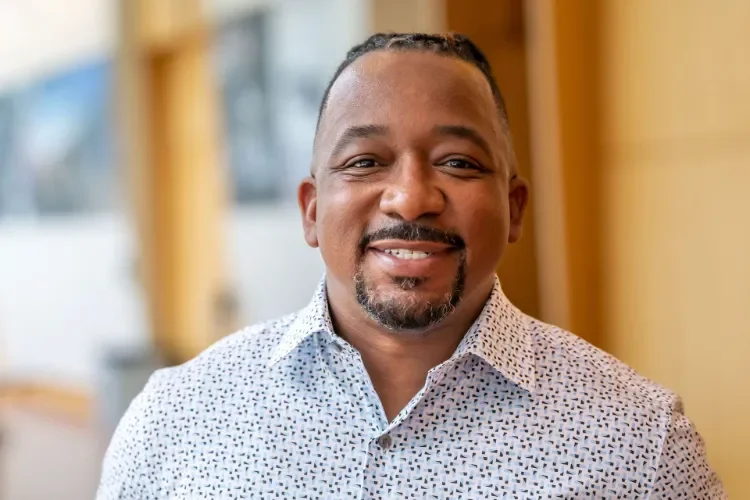 A smiling man with short hair and a goatee, wearing a white patterned shirt, indoors with a blurred background of windows and wooden elements.