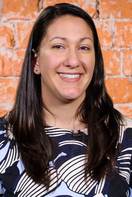 A woman with long dark hair, smiling, standing in front of a brick wall, wearing a patterned top.