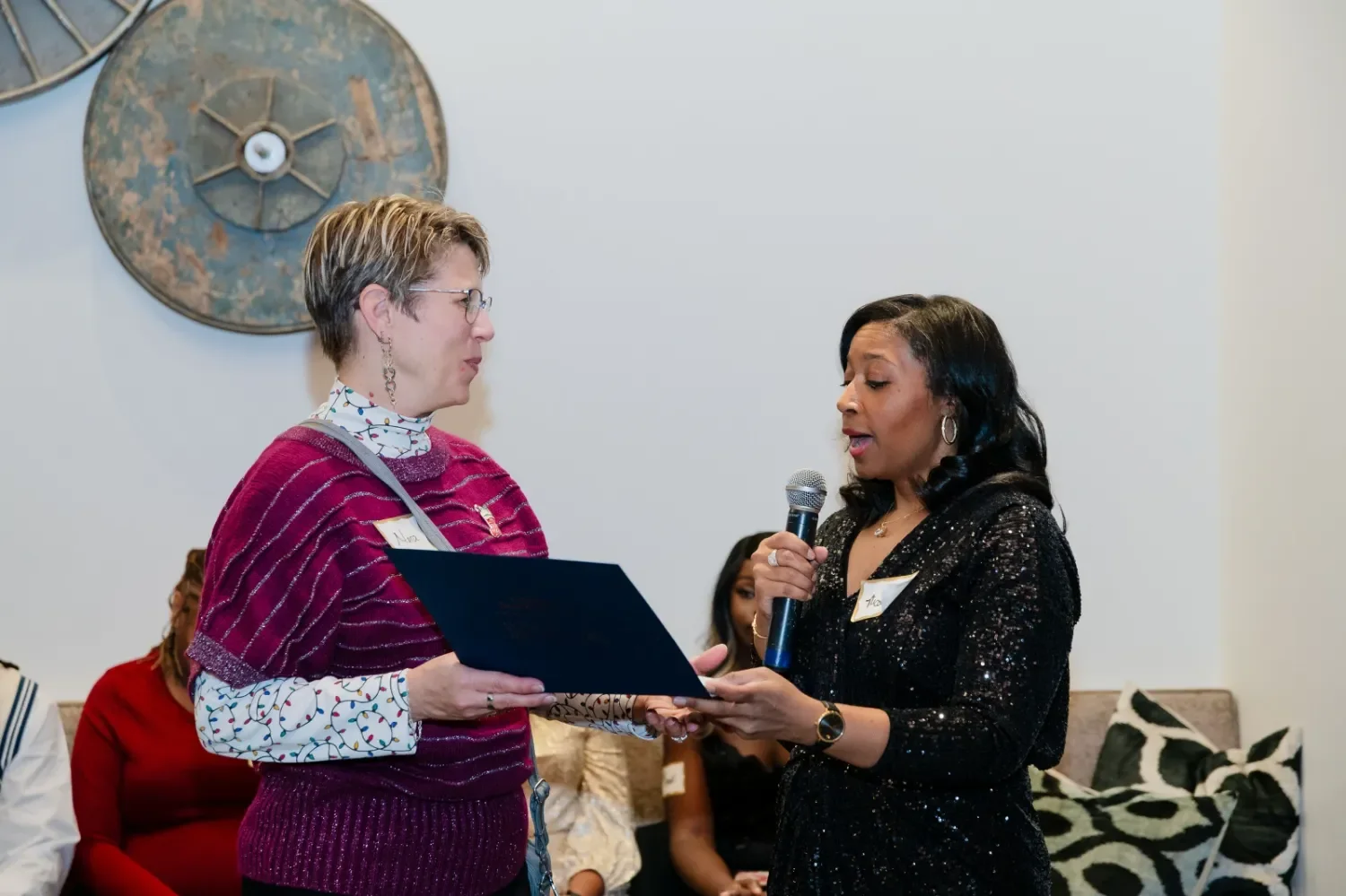 Two women are engaged in a conversation, with one woman holding a microphone and the other holding a document. They appear to be at a formal event, with a group of people seated in the background.