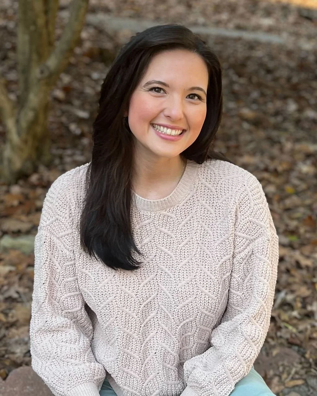 A woman with long dark hair smiling outdoors in a fall setting, wearing a light pink knitted sweater.