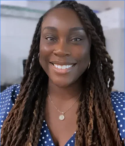 A woman with long dreadlocks wearing a blue polka dot shirt and a necklace, smiling at the camera.