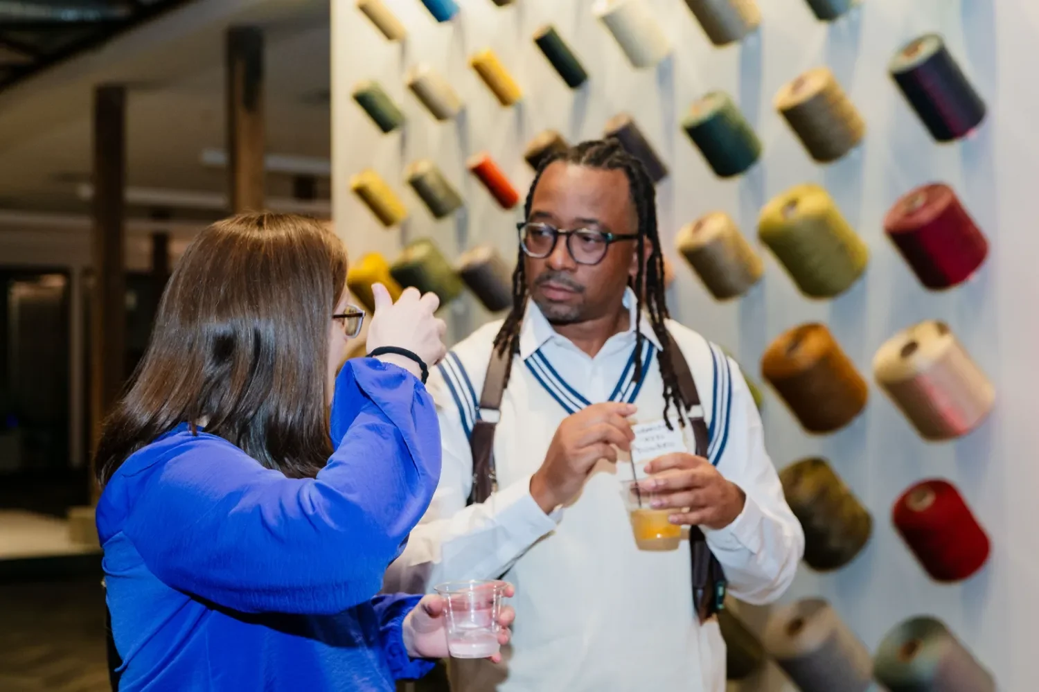 Two people, a woman in a blue jacket and a man with glasses and dreadlocks, are talking and holding drinks in front of a wall display of colorful spools of yarn.