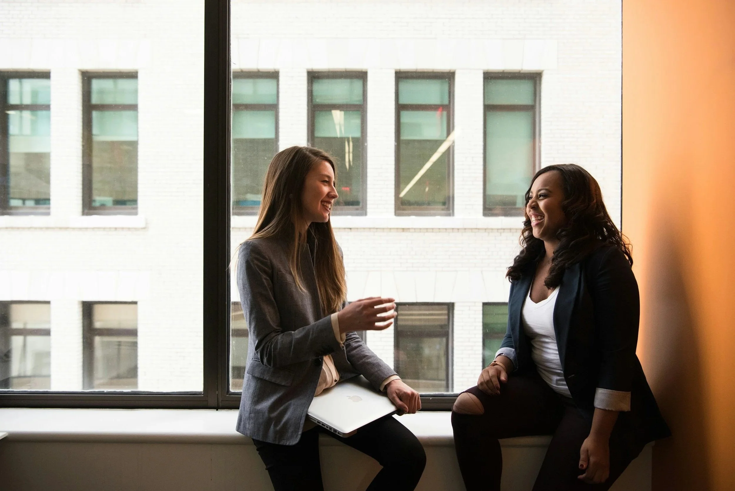 Two women are sitting inside by a large window, engaged in conversation and smiling. One is holding a laptop.