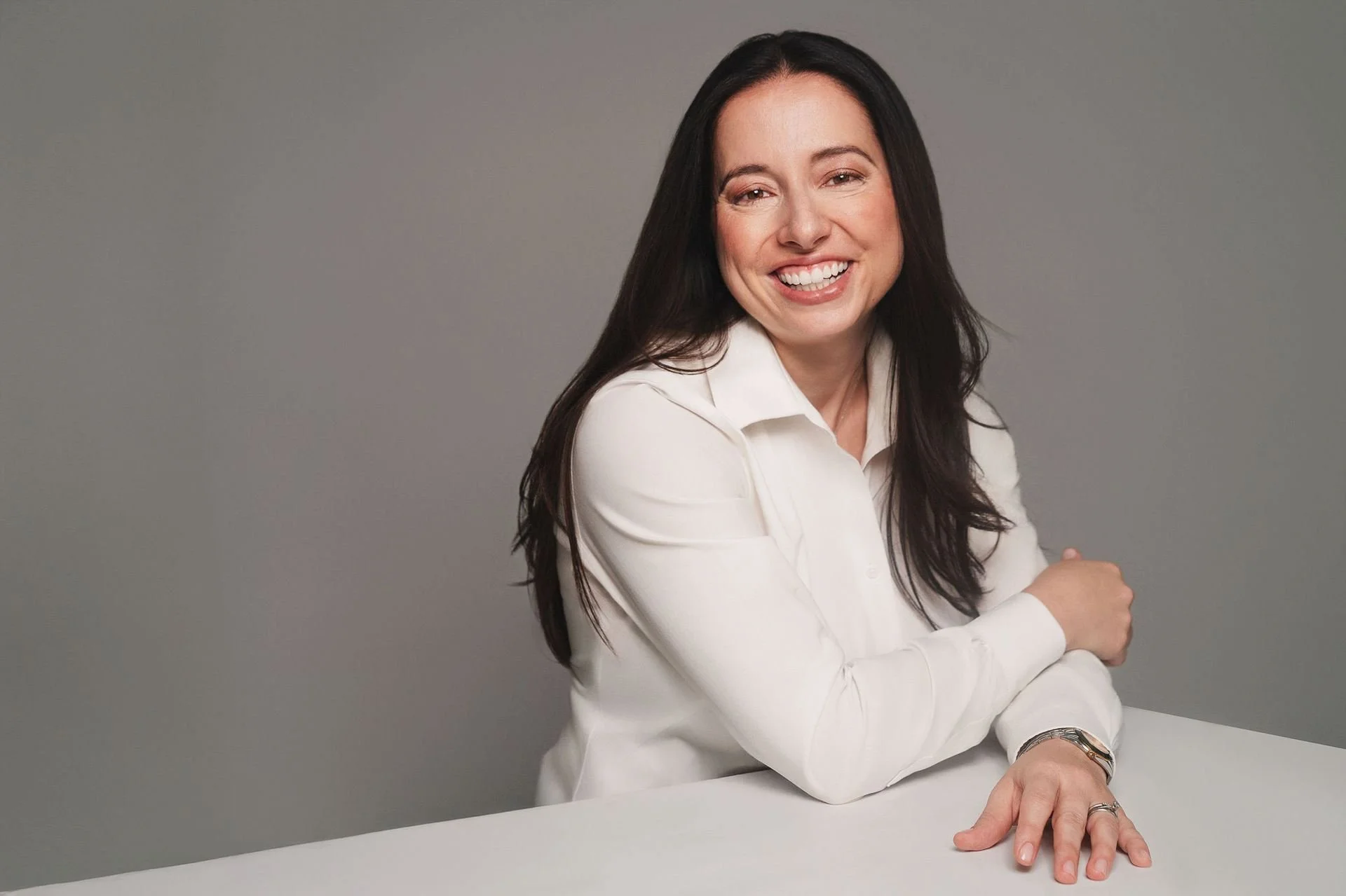 A woman with long dark hair, smiling, wearing a white blouse, sitting at a table against a gray background.