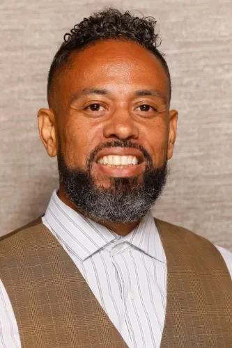 A smiling man with curly black hair and a full beard, wearing a white striped shirt and a tan vest, against a neutral background.