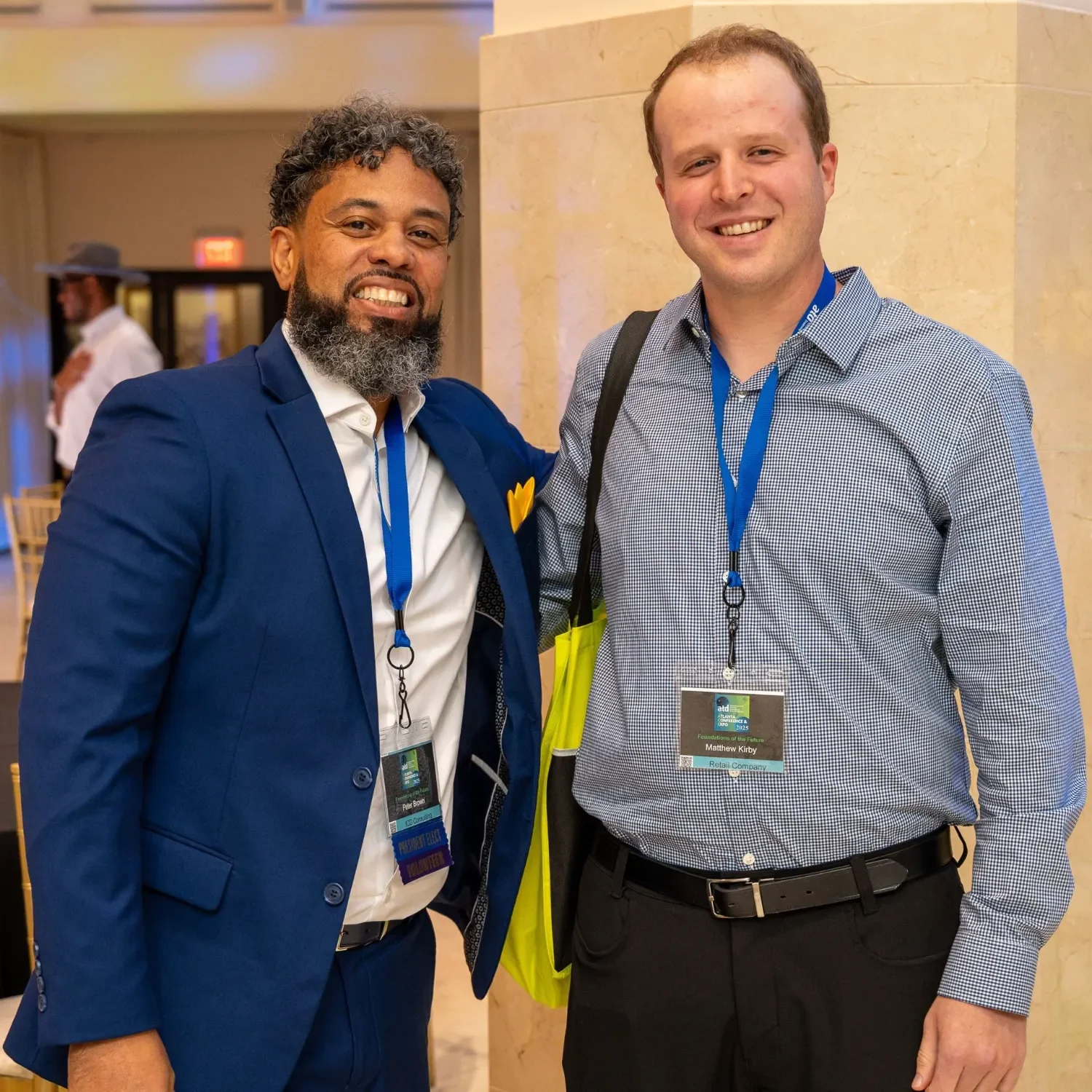 Two men smiling at an indoor event, wearing conference badges and business attire, with a man in the background adjusting his suit