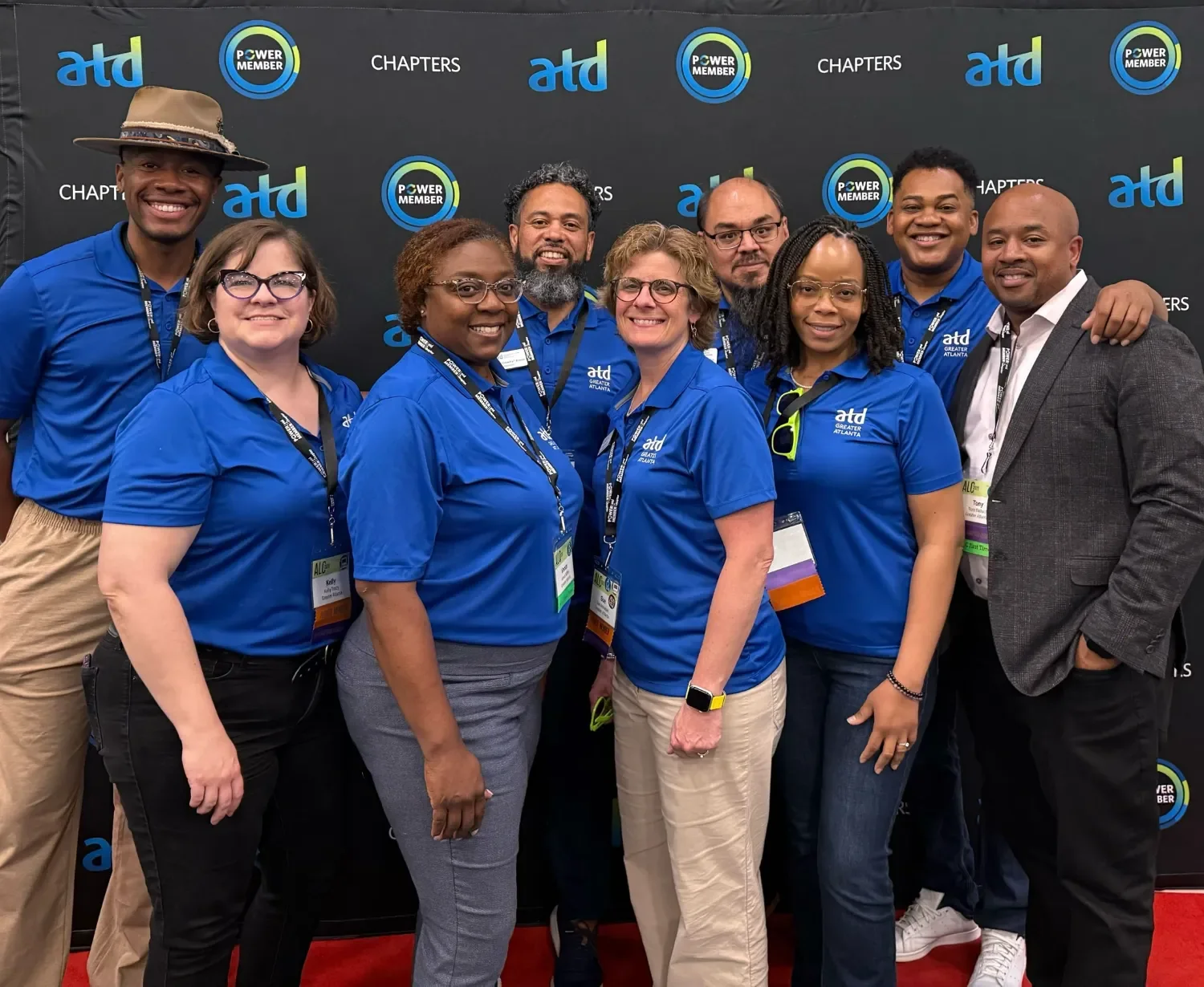 Group of nine diverse individuals smiling and posing together at an event, all wearing blue shirts with 'ATD Greater Atlanta' logos, standing in front of a black backdrop with the logos 'atd' and 'Power Member' repeatedly printed on it.