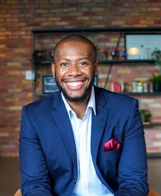A smiling man in a blue suit with a pink pocket square, seated indoors with a brick wall and shelves in the background.