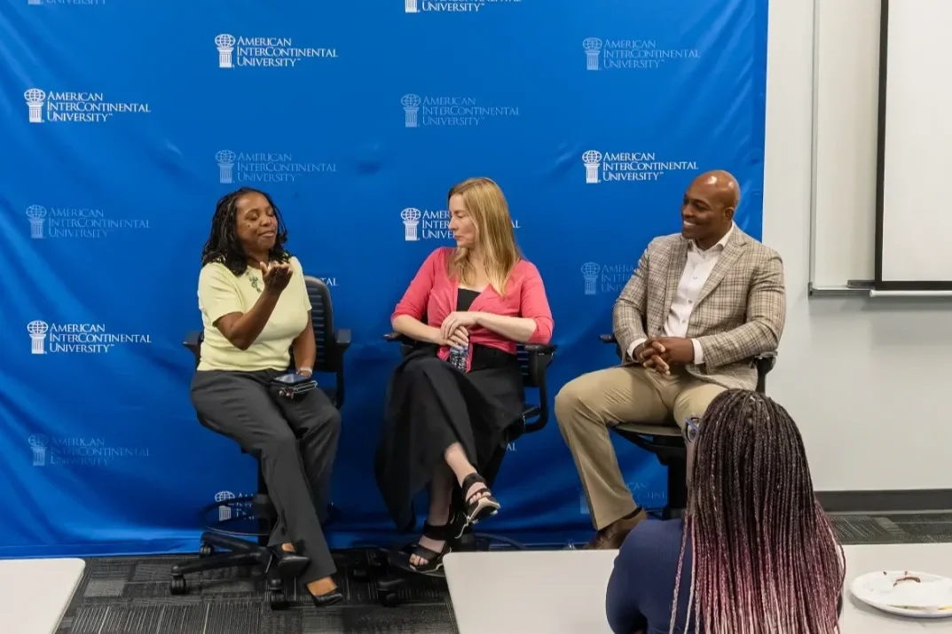Three people engaged in a panel discussion or interview in front of a blue backdrop with the American Intercontinental University logo, seated on chairs, with an audience member visible in the foreground.