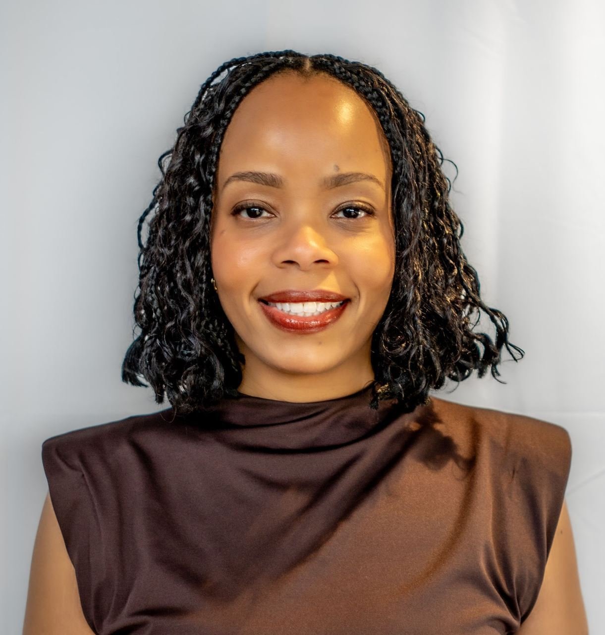 A portrait of a smiling woman with curly black hair, wearing a dark brown sleeveless top, standing against a light-colored background.