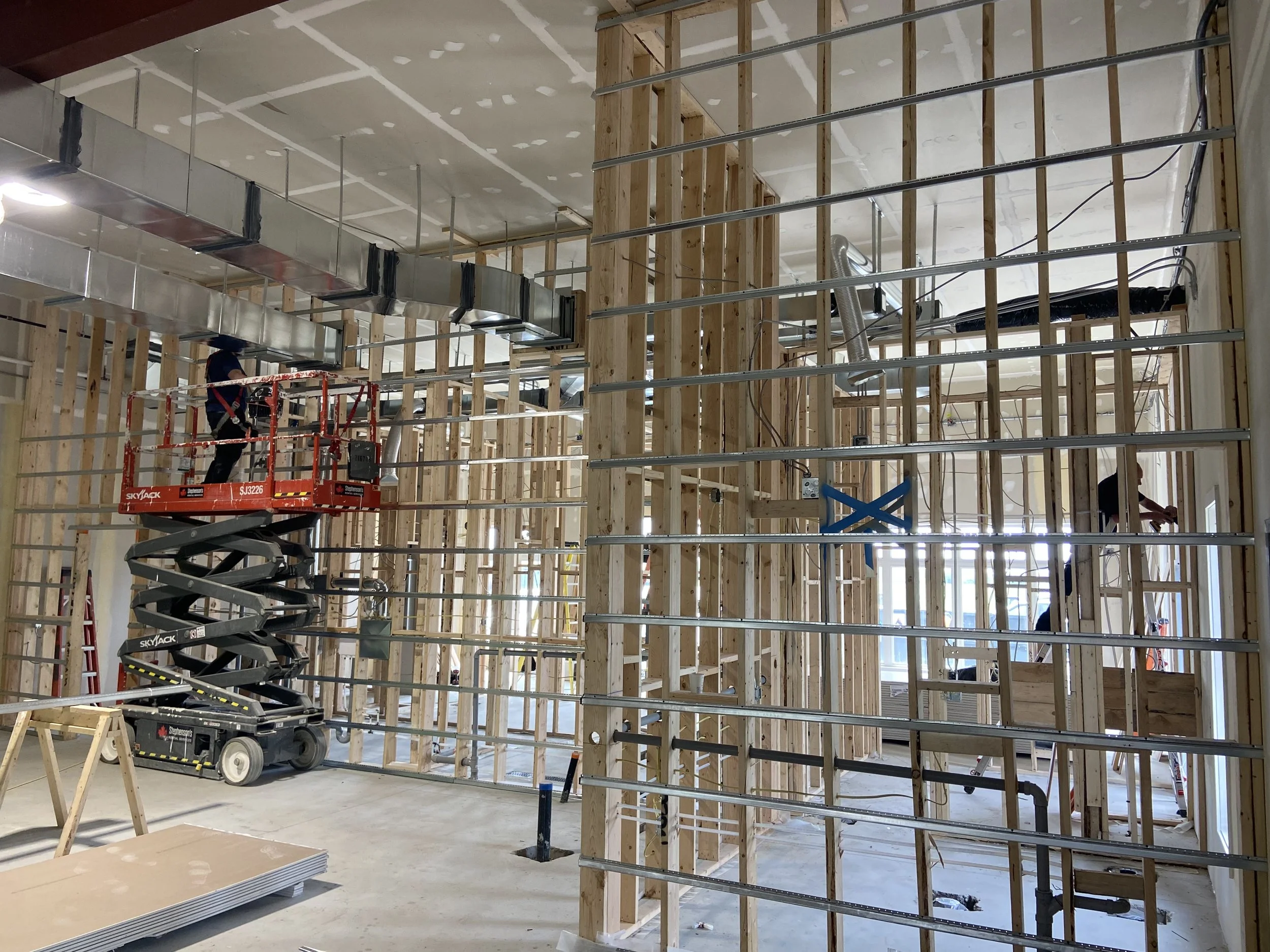 Construction workers install ductwork and electrical wiring inside a building's framed interior. The room has exposed wooden studs and ceiling, with a lift and scaffold present.