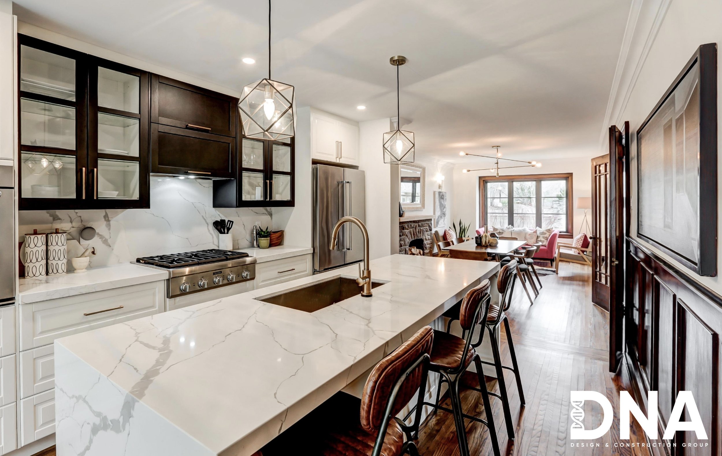 Modern kitchen with white marble island and dark upper cabinets, stainless steel appliances, and a view into a cozy living and dining area with large windows and seating.