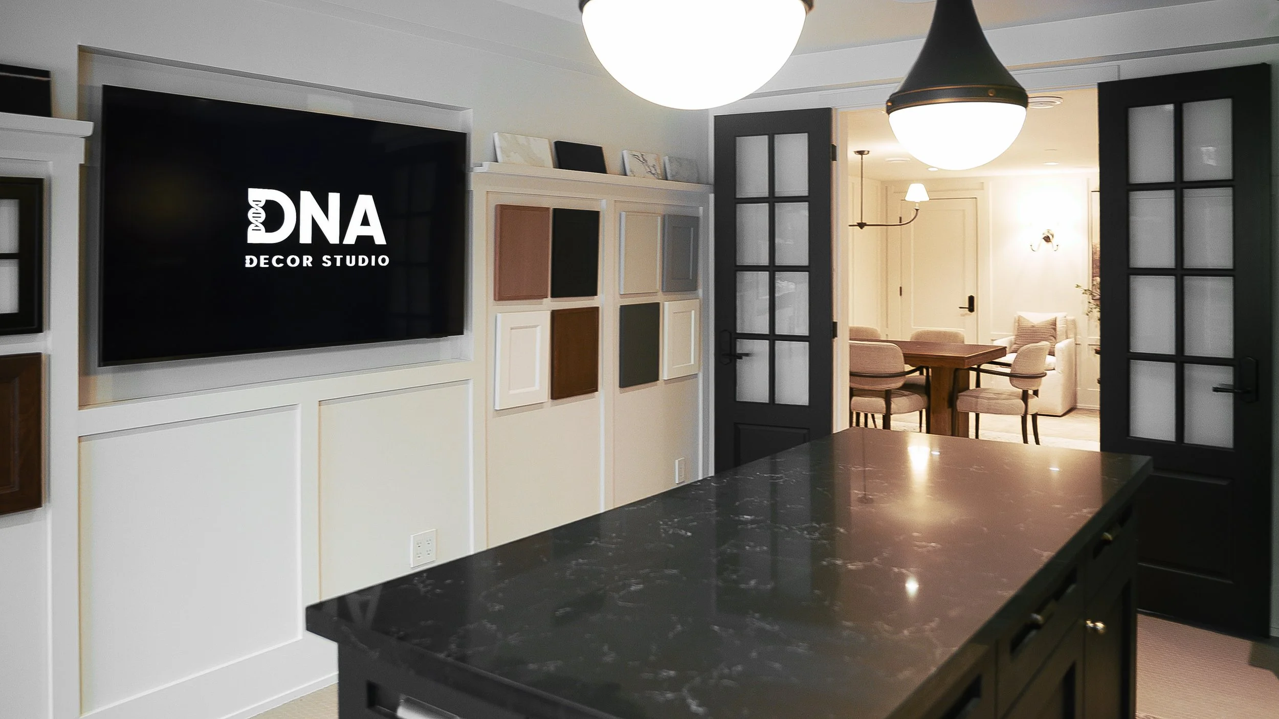 Interior of a modern living space with a large black marble kitchen island in the foreground, black-framed glass double doors open to a brightly lit dining area with a wooden table and beige chairs, a wall-mounted TV displaying 'DNA Decor Studio', an