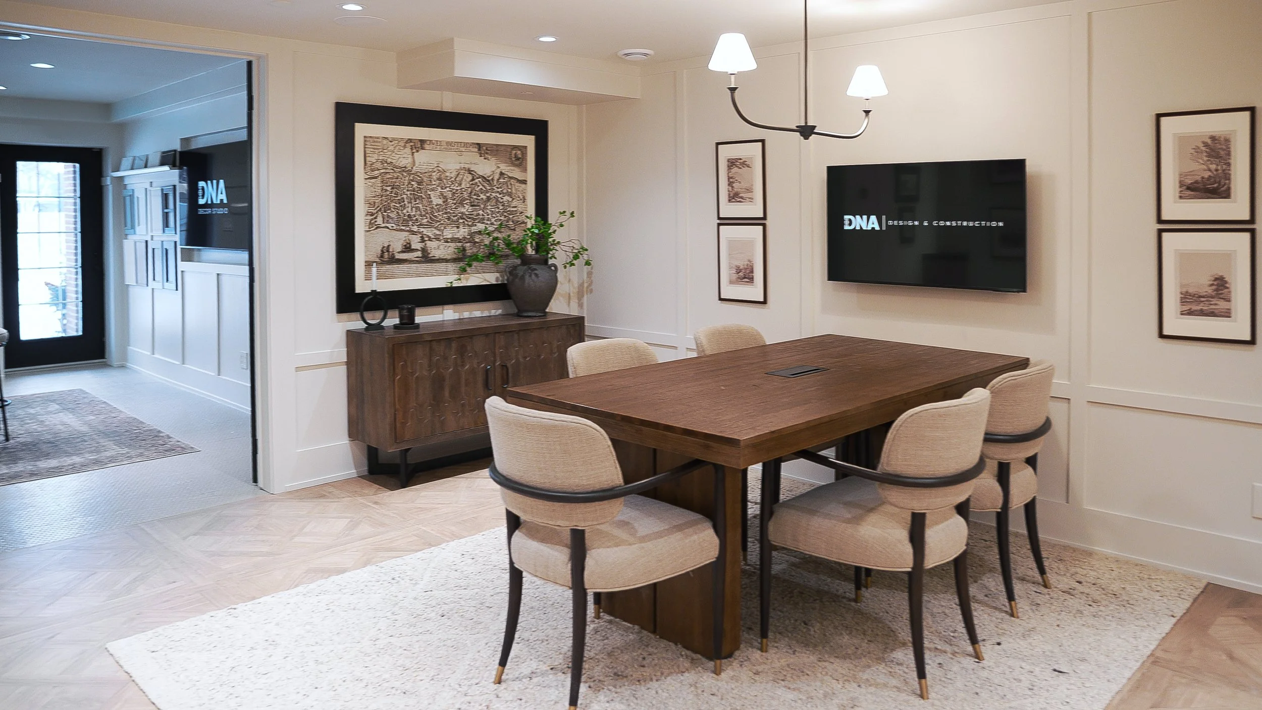 A dining room with a wooden table surrounded by beige upholstered chairs, a wall-mounted television, framed artwork, a sideboard with decorative items, a large potted plant, and a chandelier with two shades.