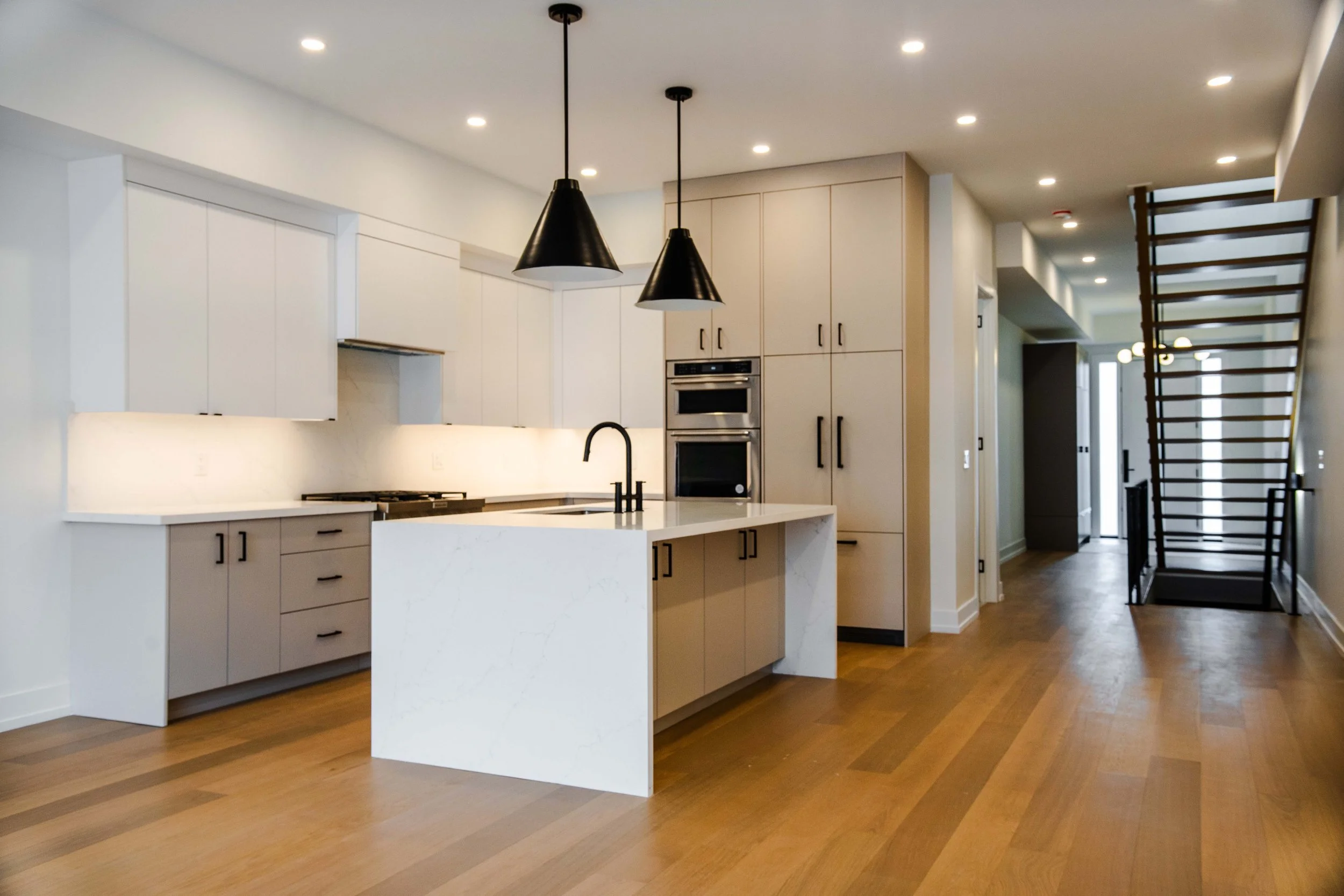 Modern white kitchen with black fixtures, wood flooring, and a staircase in the background.