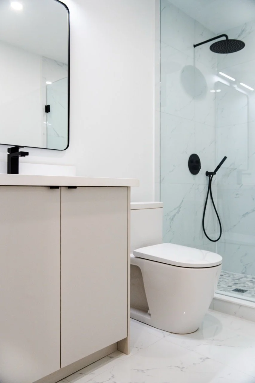 Modern bathroom with white vanity, black faucet, mirror, toilet, and walk-in shower with black fixtures and marble walls.