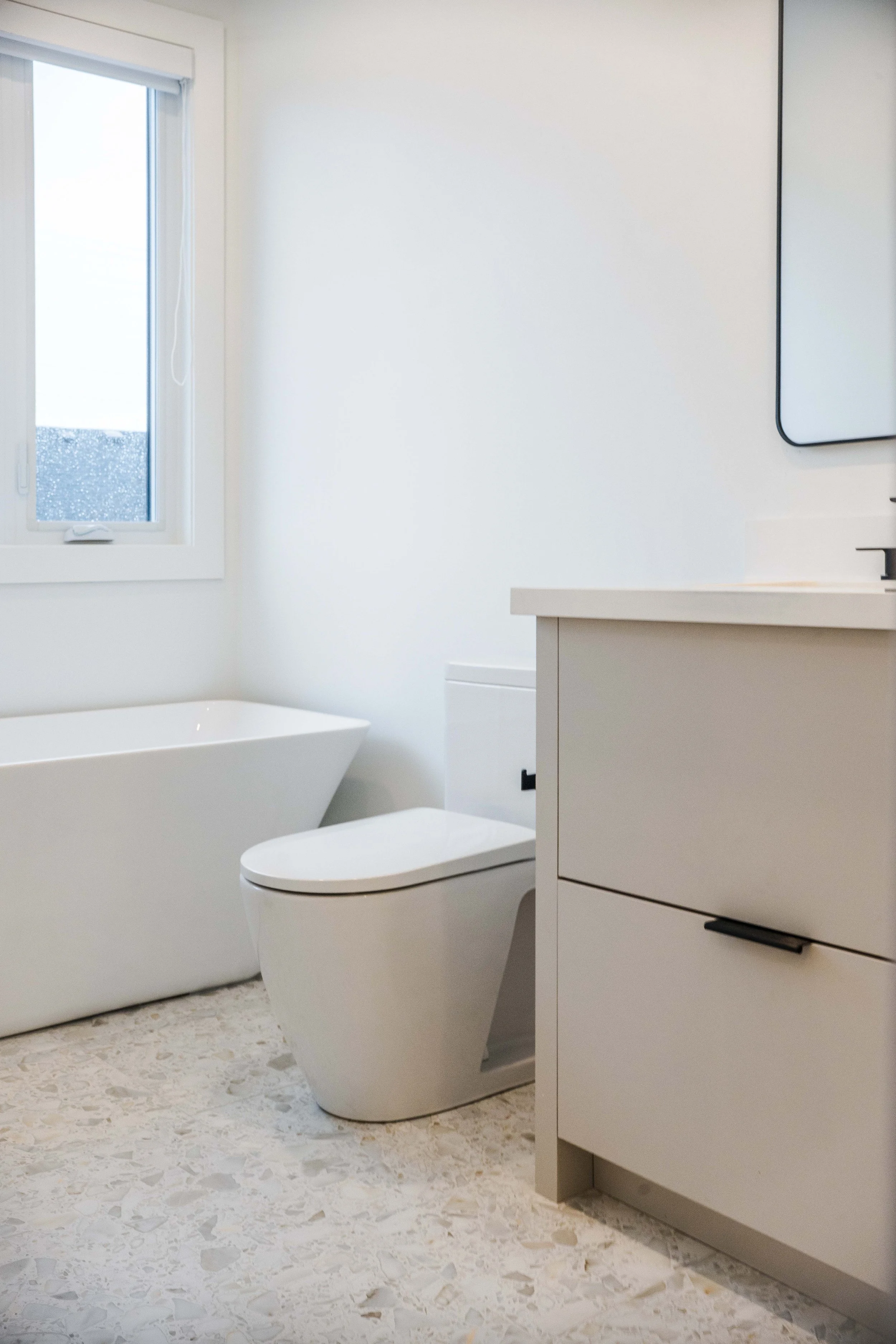 Modern bathroom with a bathtub, toilet, and white vanity with black handles, natural light from a window, light-colored terrazzo flooring.