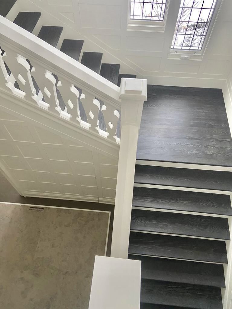 Interior view of modern staircase with black steps and white balustrade, viewed from the top, looking downward towards a window with grid pattern, natural light coming through.
