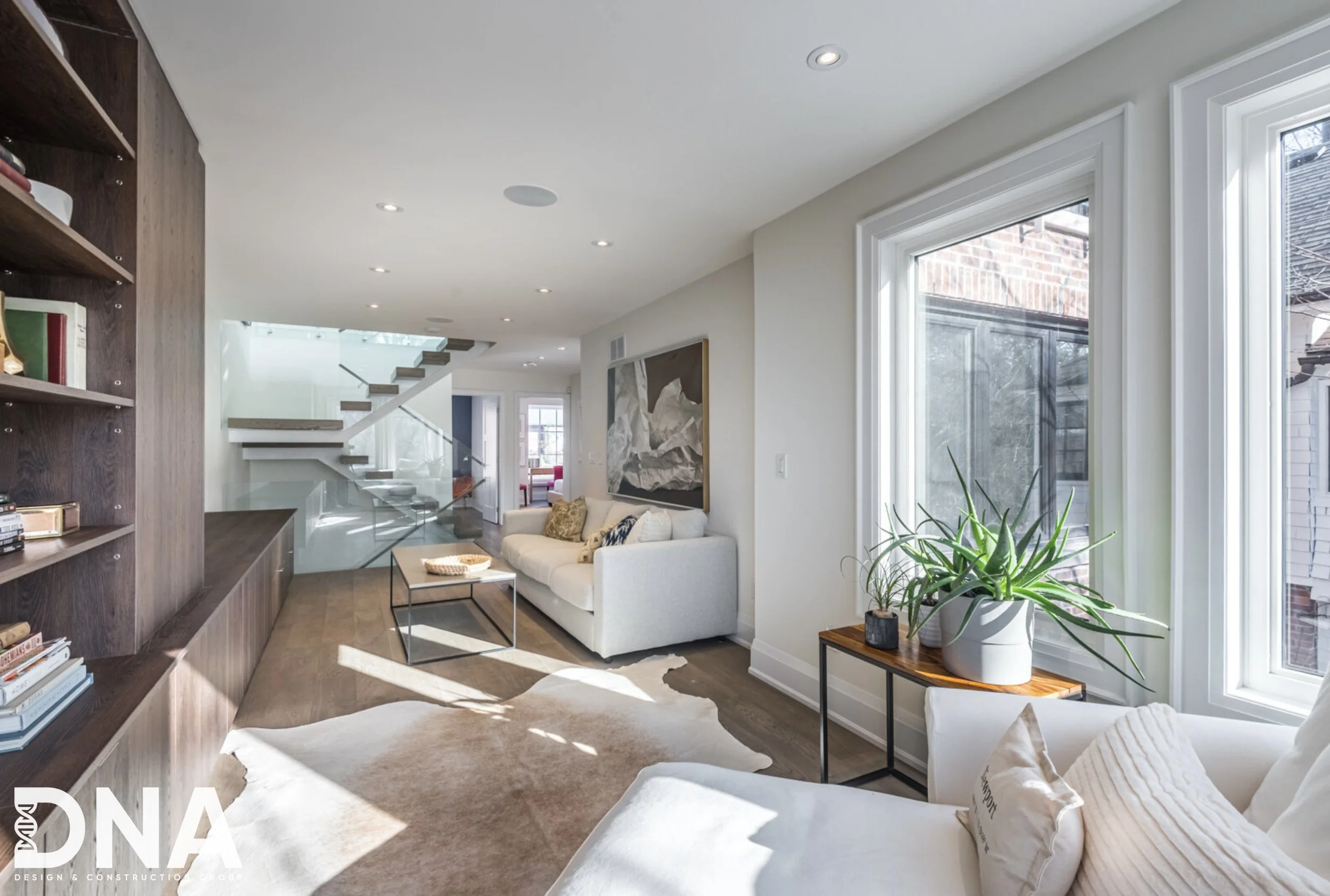 Modern living room with white sofas, wooden side table with plants, bookshelf, large windows, artwork, staircase with glass railing, and sunlight streaming in.