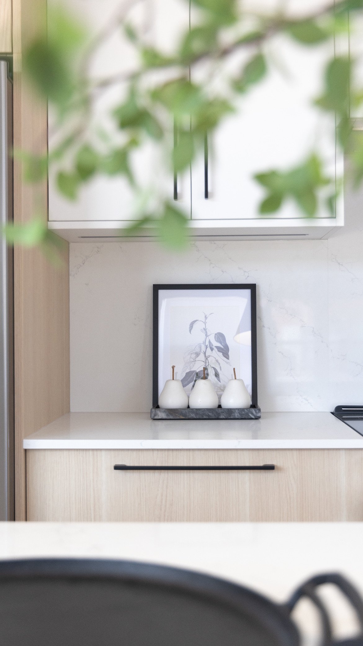 Kitchen countertop with three white ceramic pears, framed botanical print, and marble tray, partially obscured by green leaves in the foreground.