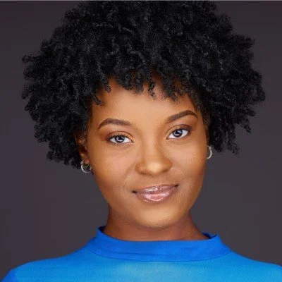 Portrait of a woman with short curly black hair wearing a blue top and small earrings, smiling softly.