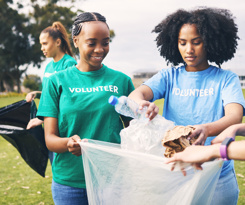 Two female volunteers collecting trash in a park, wearing teal and blue shirts that say 'Volunteer', with a third person in the background also picking up litter.