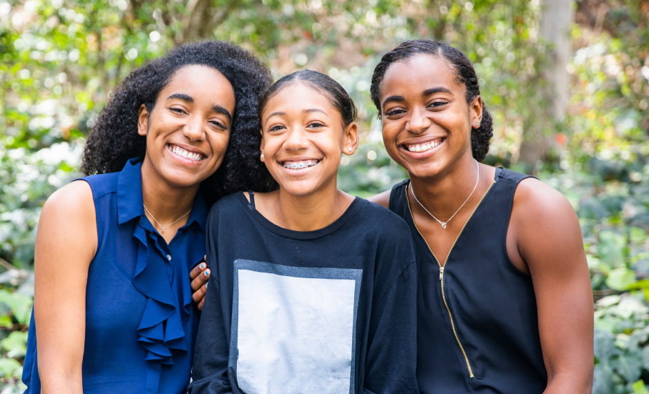 Three young women smiling outdoors in a green, natural setting.