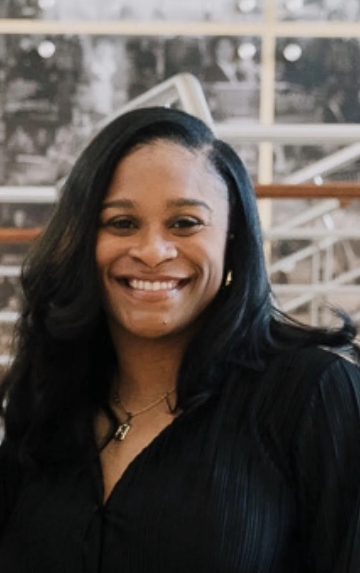 A smiling woman with long black hair, wearing a black top and a necklace, standing indoors with a blurred background.