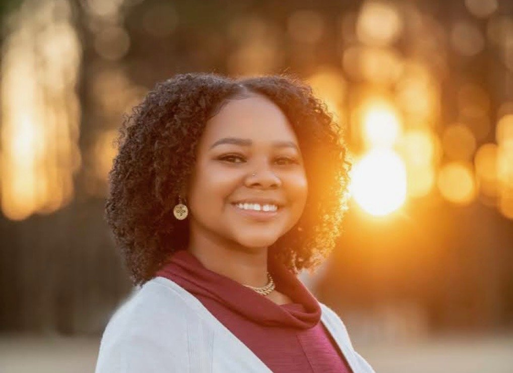 A woman with curly hair smiling outdoors during sunset, wearing earrings, a necklace, a maroon top, and a light jacket.