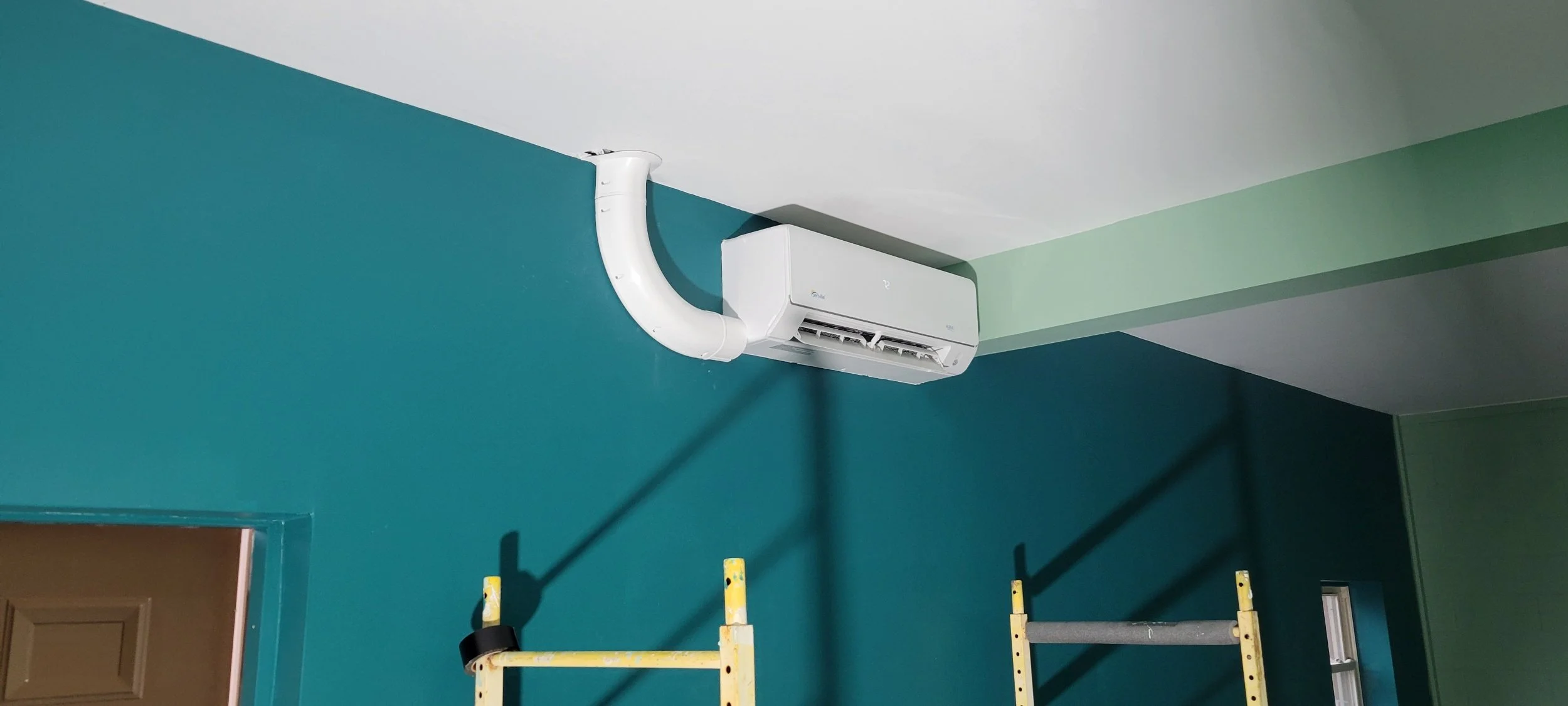 A wall-mounted air conditioning unit installed high on a teal-colored wall near the ceiling, with a white vent pipe running upward and a yellow step ladder in front of the wall.