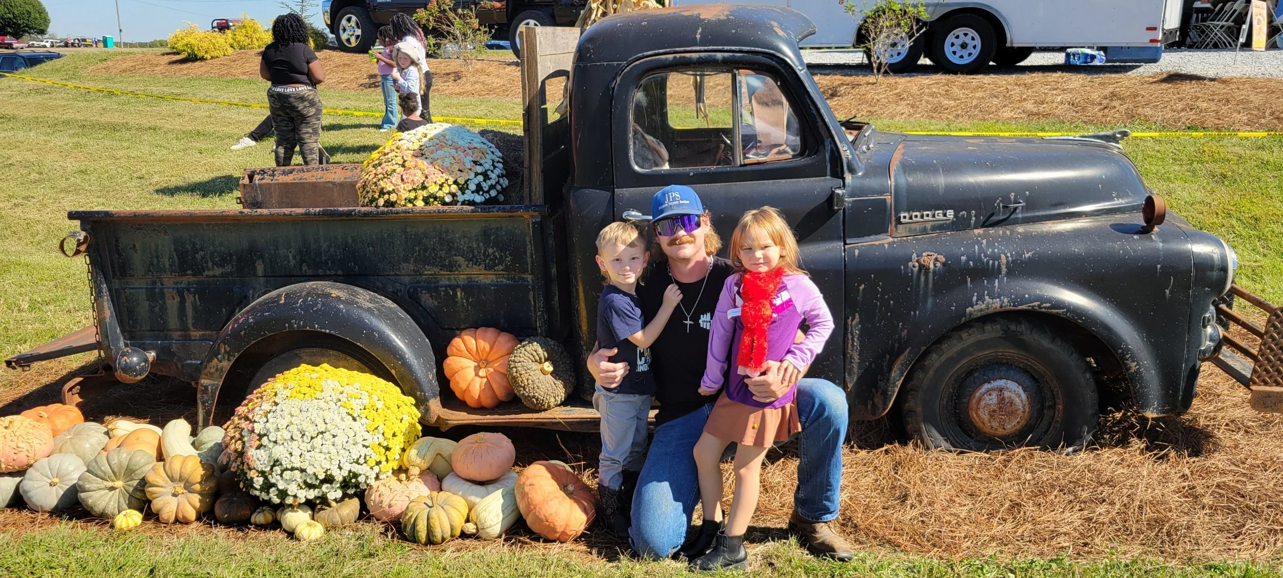 A vintage black pickup truck decorated with pumpkins and fall flowers, with three children and an adult posing in front of it at an outdoor fall event.