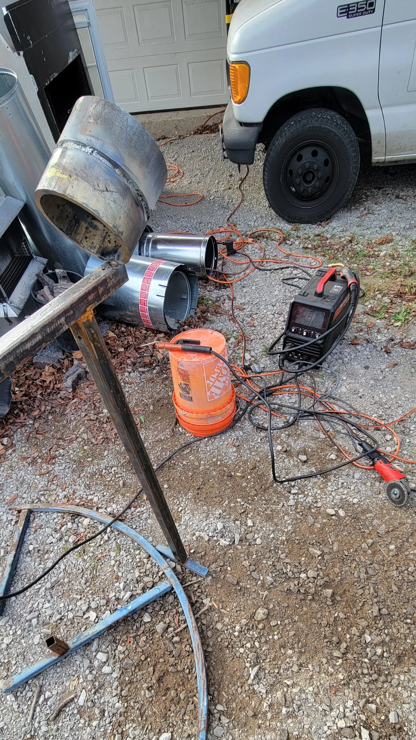 Garage with welding equipment, extension cords, and metal parts on gravel driveway.