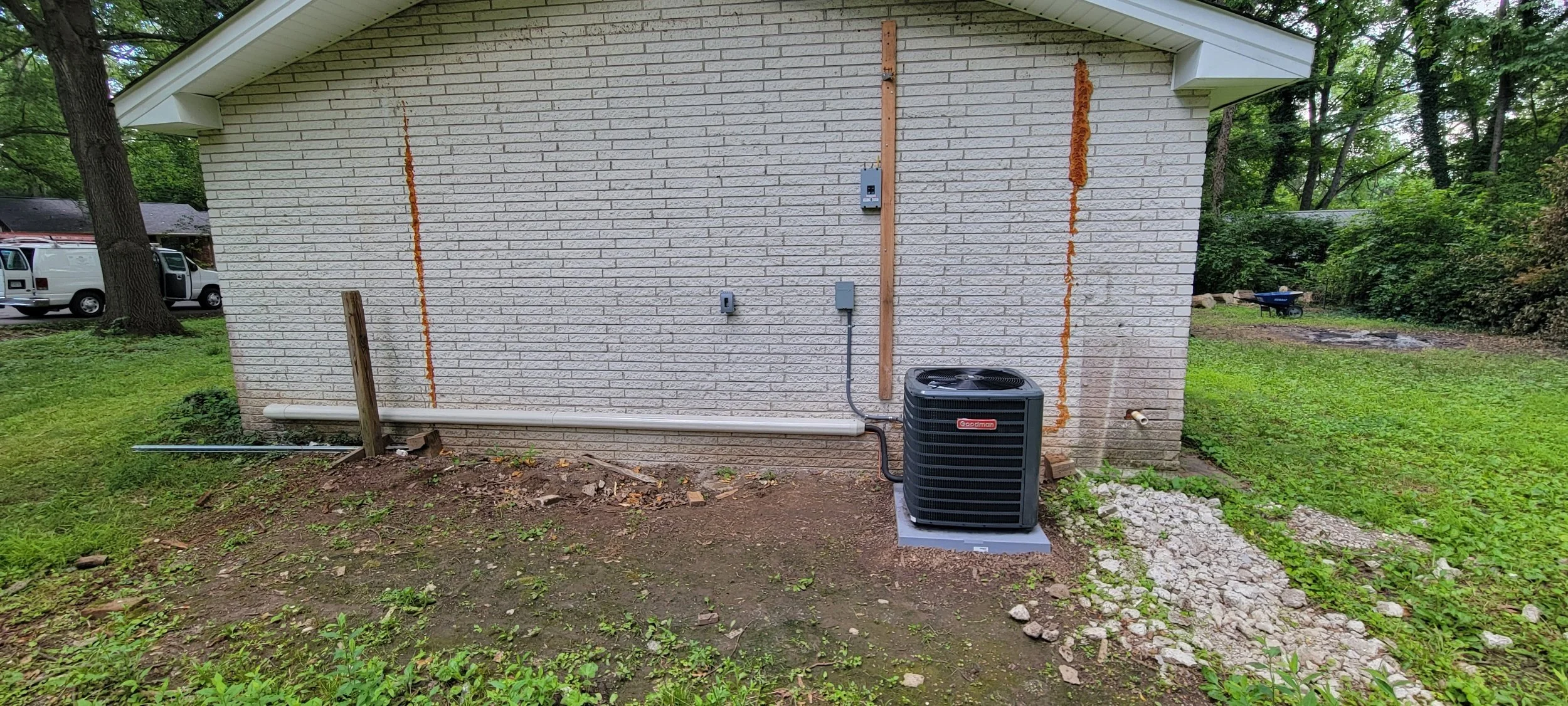 Backyard with white brick house wall, outdoor HVAC unit, pipes, and electrical boxes, surrounded by green grass and trees.