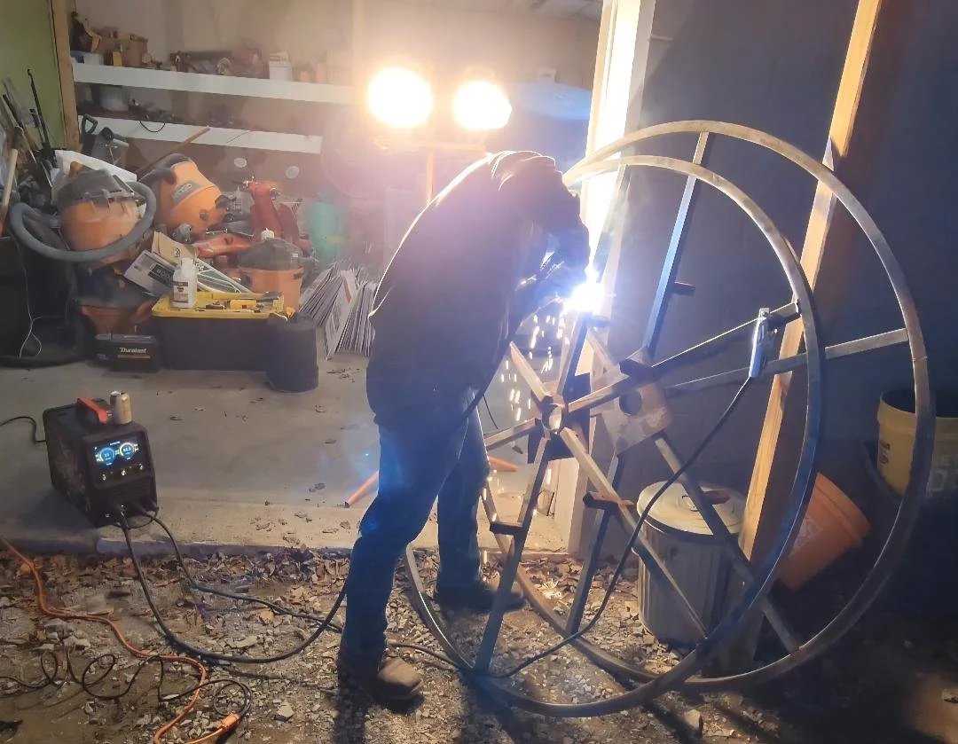 A person welding a large metal circular structure inside a garage or workshop. Sparks are flying from the welding process, and tools and equipment are scattered around.