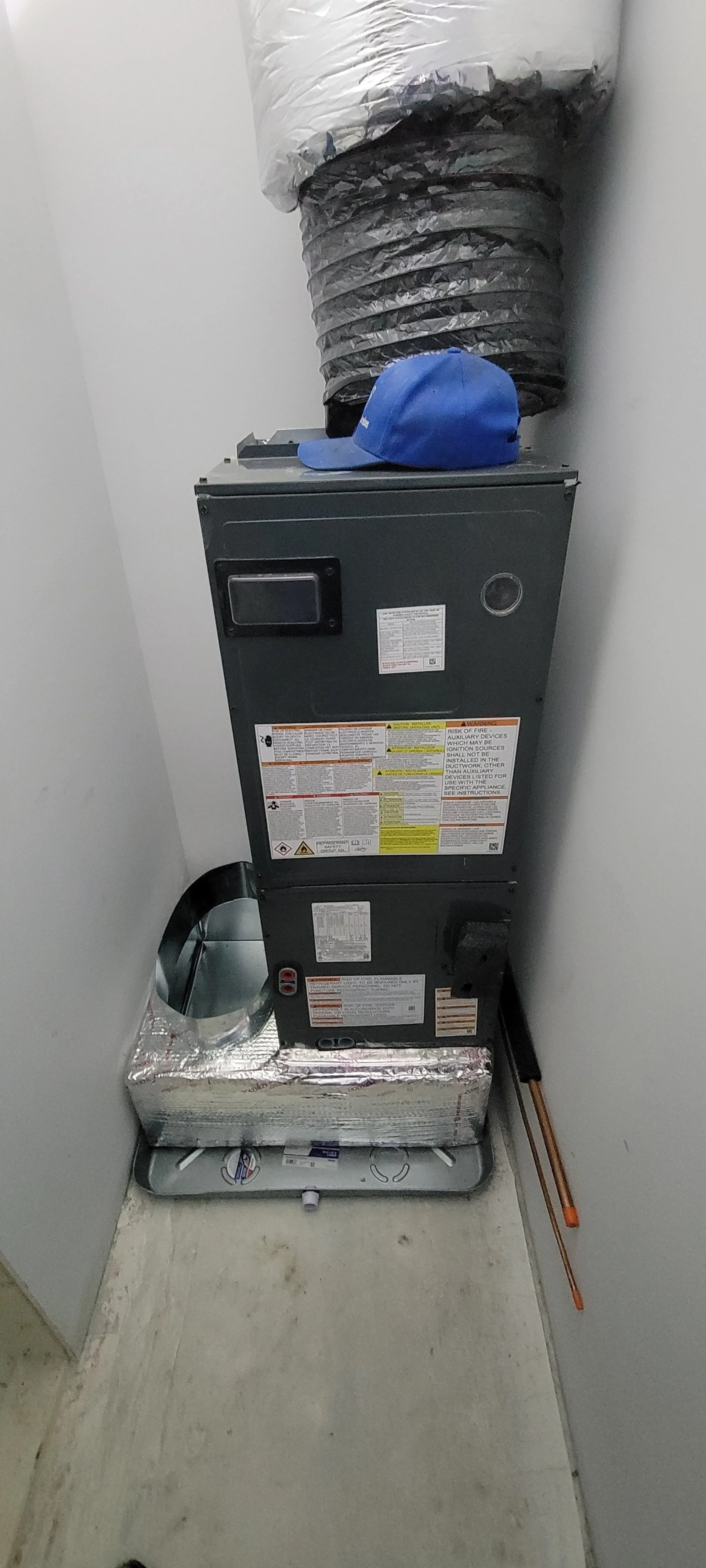 A furnace with a blue cap placed on top, a large black and silver flexible duct connected to it, and a flexible metal pipe on the floor in a narrow space between two white walls.