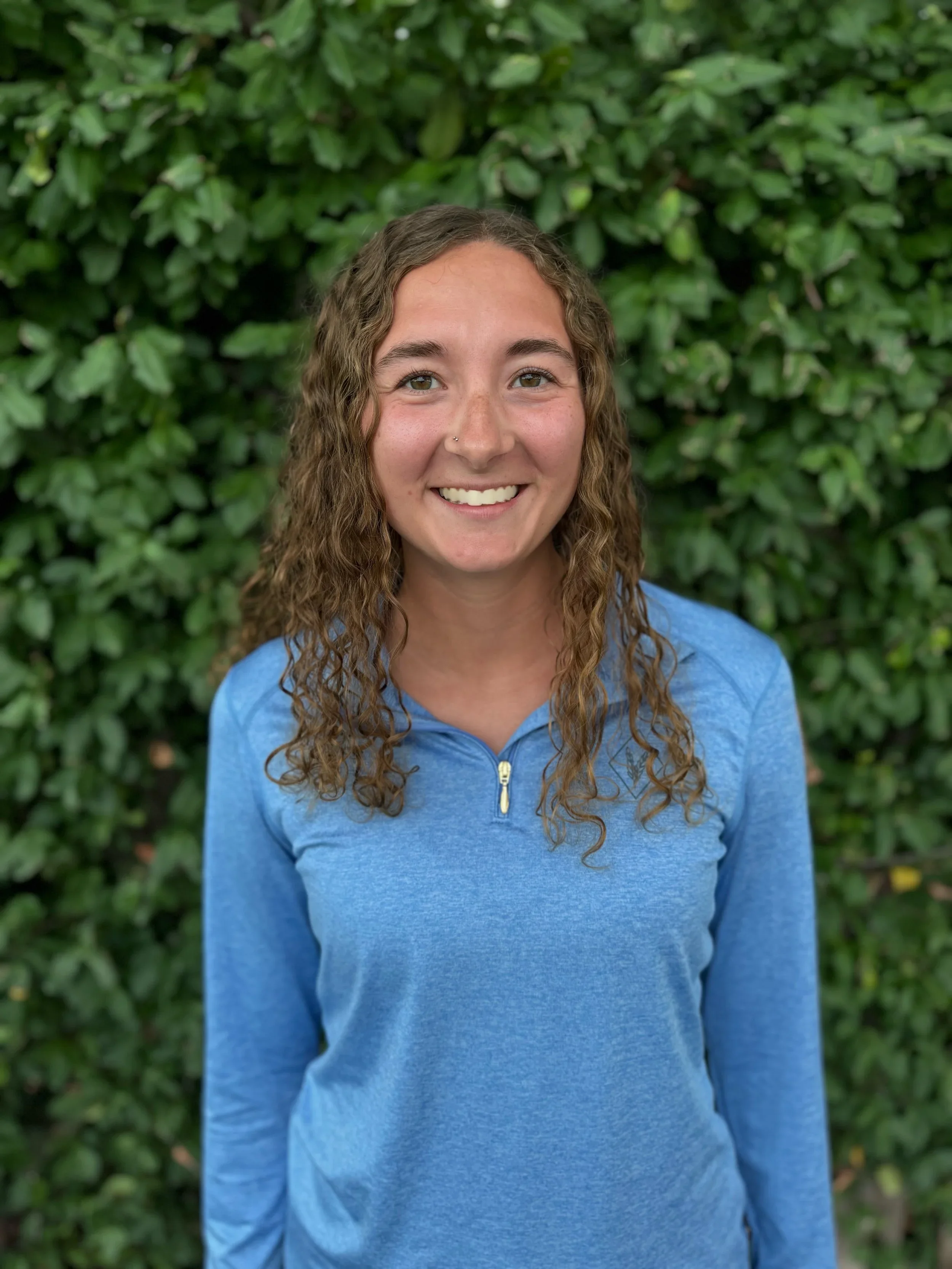 A woman with curly brown hair, smiling, wearing a blue quarter-zip top, standing outdoors in front of a green leafy background.