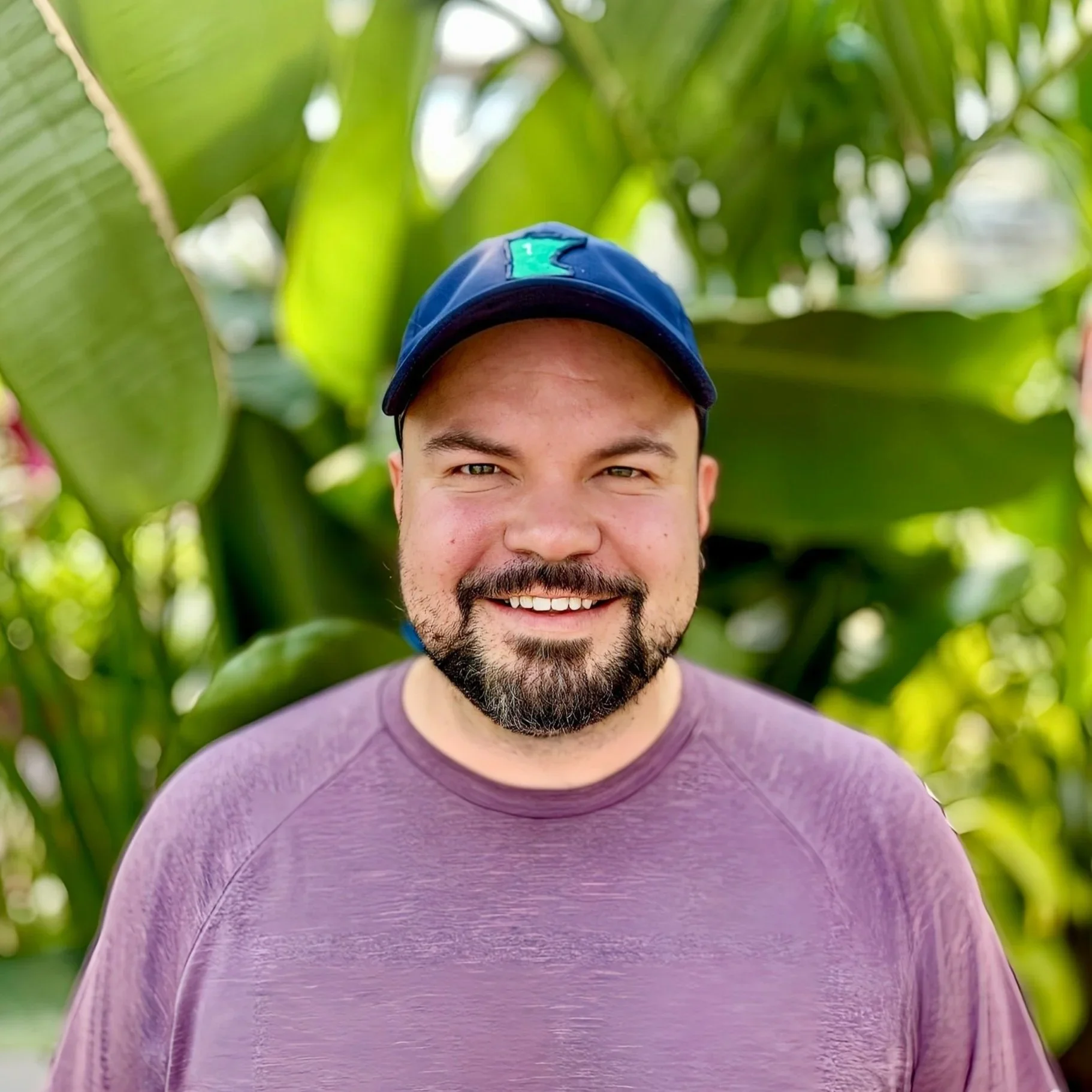 A smiling man with a beard and mustache wearing a purple shirt and a blue cap, standing in front of lush green tropical plants.