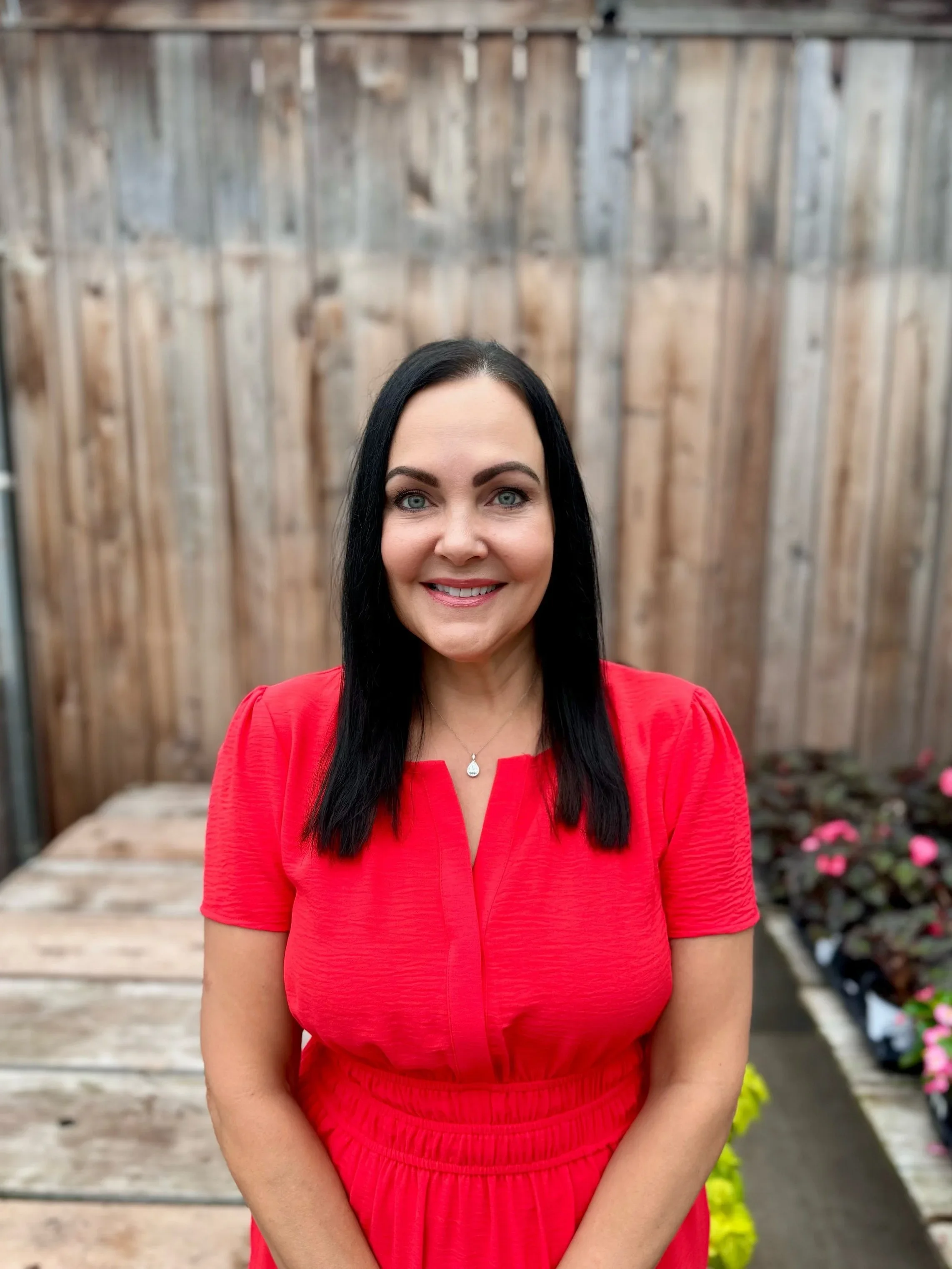 A woman with black hair and light skin, smiling, wearing a red dress, standing outdoors with a wooden fence and potted flowering plants in the background.