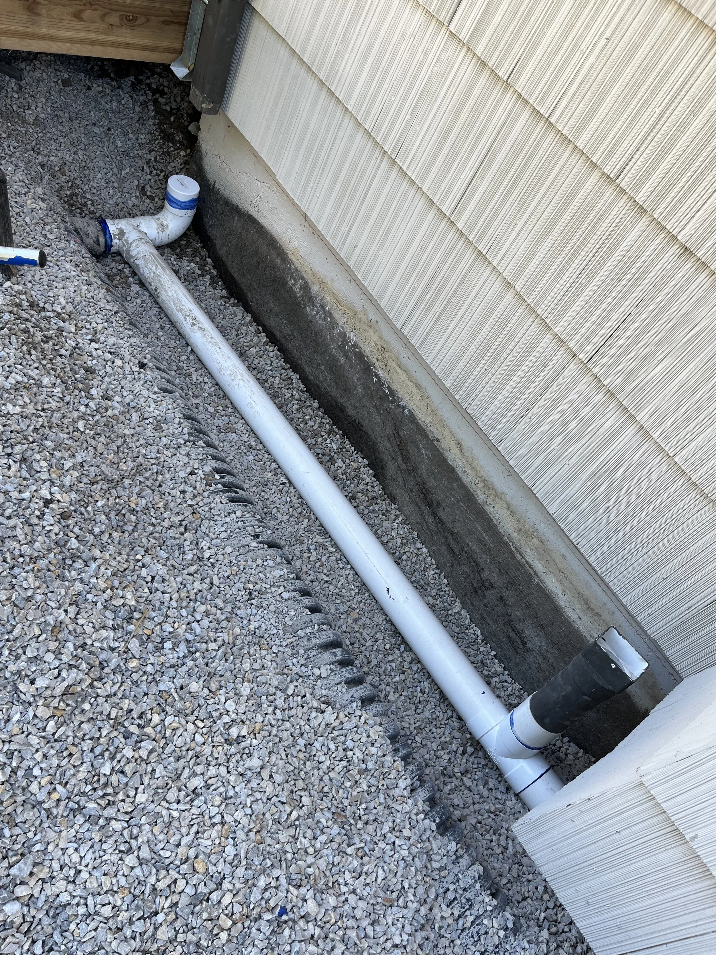 PVC pipes installed near the foundation of a house, with gravel on the ground and a downspout connection at the corner of the house, for drainage or plumbing system.